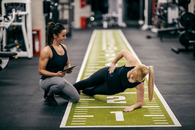 A sportswoman is doing side planks while her female personal trainer is watching her