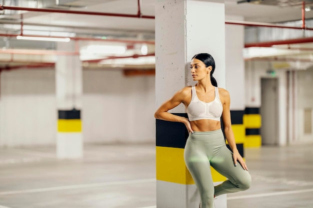 A sportswoman is leaning on the pillar in underground garage and posing