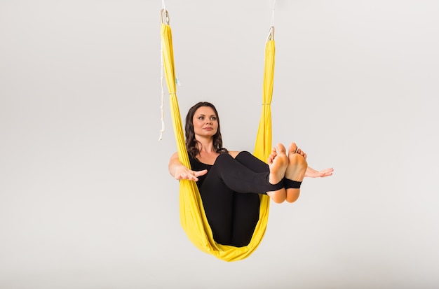 Sportswoman performs the Paripurna Navasana pose in a yellow hammock on a white wall