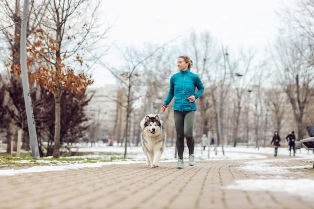 Sportswoman running together with her dog in park on snowy weather. Winter fitness, pets, friendship