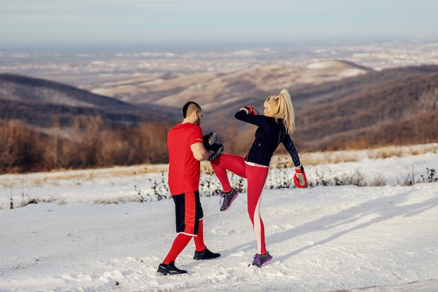 Sportiva sparring e calci pad in natura al giorno d'inverno nevoso con il suo allenatore. boxe, fitness invernale, fitness all'aperto