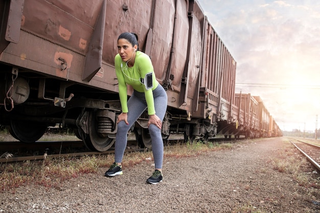 Sportswoman with earphones taking a break after running by trains at the station.