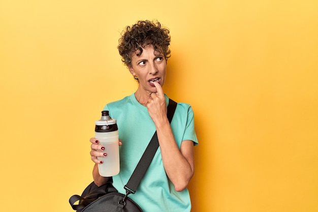 Sportswoman with gym bag water on yellow studio relaxed thinking about something