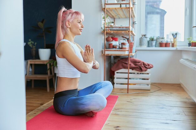 Sportswoman with ponytail holds hands in namaste sitting in lotus pose indoors