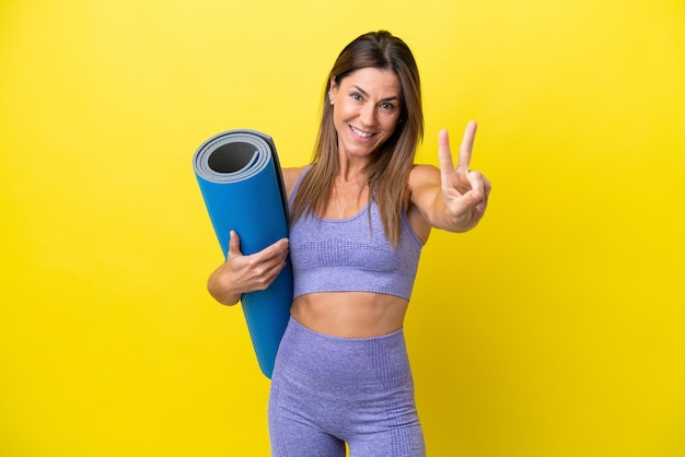 Sportvrouw die naar yogalessen gaat terwijl ze een mat geïsoleerde niet-gele achtergrond vasthoudt die lacht en een overwinningsteken toont