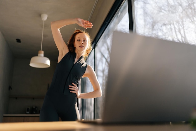 Sportvrouw die oefent, zich opwarmt, fitnessvideo's online kijkt op laptop thuis.