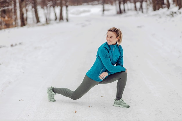 Sportvrouw gehurkt in de natuur op sneeuw in de winter en opwarmingsoefeningen doen. Natuur, bos, winterfitness, gezonde levensstijl