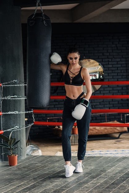 Sportvrouw in zwarte sportkleding en witte bokshandschoenen rust na een zware training in de moderne loft gym. Sportieve levensstijl concept.