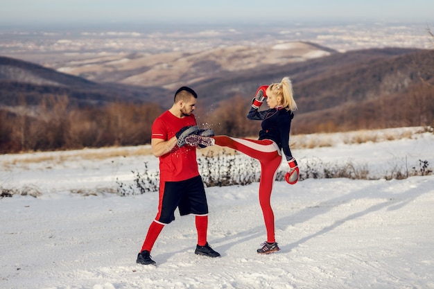 Sportvrouw sparren met bokshandschoenen in de natuur op besneeuwde winterdag met haar instructeur. Ze is aan het schoppen. Boksen, winterfitness, buitenfitness