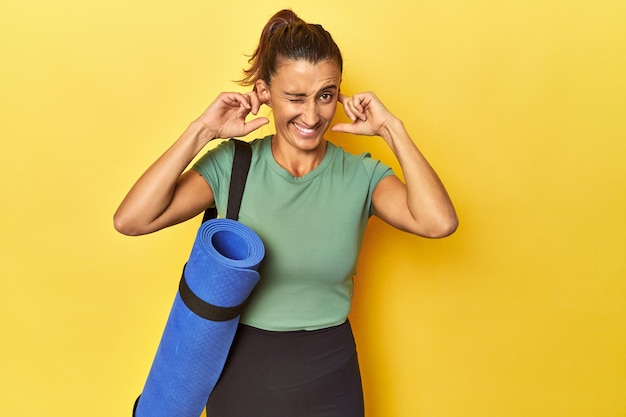 Sportvrouw van middelbare leeftijd met yogamat op gele studio die oren bedekt met handen
