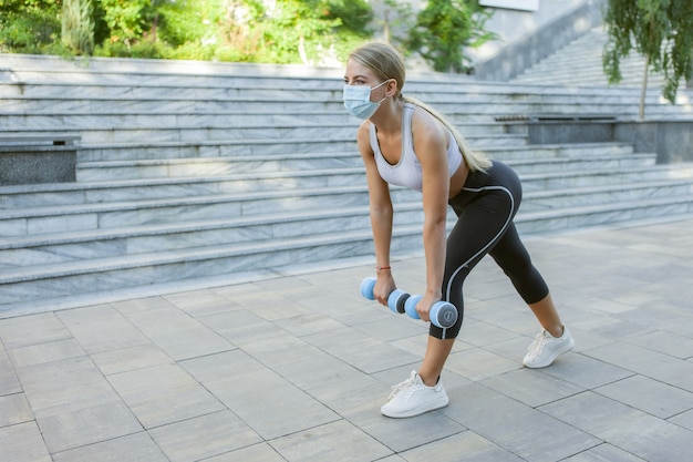 Sportwoman in abiti sportivi ed esercizi di maschera protettiva medica con manubri all'aperto. lezioni di fitness durante il covid-19