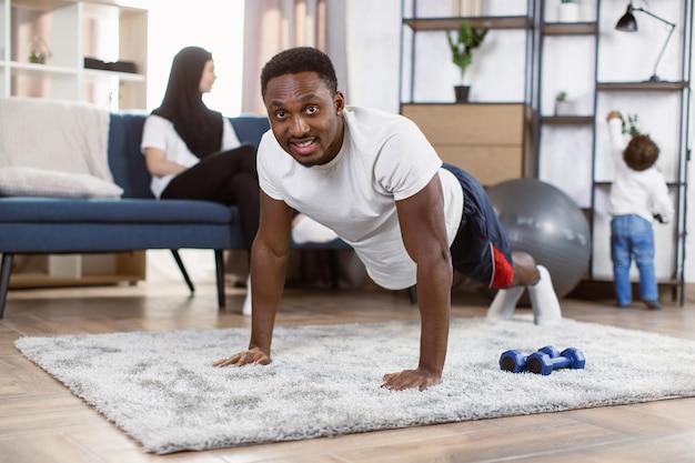 Sporty african man doing yoga plank looking at camera with muslim wife and son on background