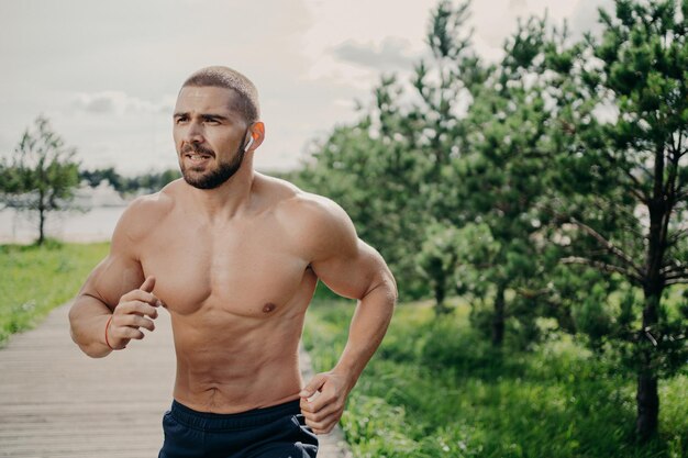 Sporty athletic man runs outdoor with naked torso leads healthy lifestyle has morning workout listens music in wireless earphones has muscular body breathes fresh air Health activities