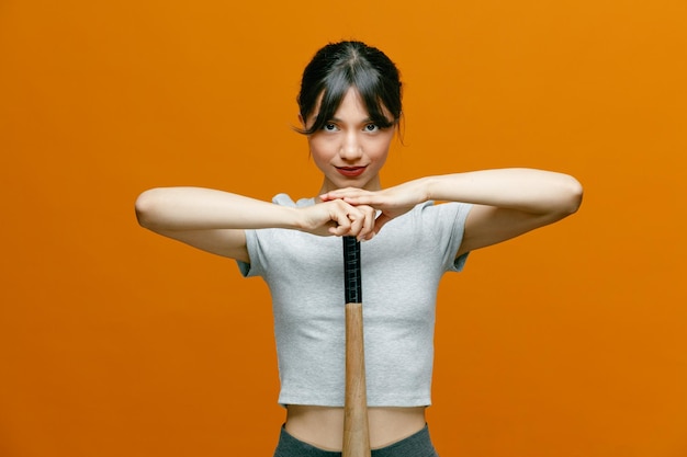 Sporty beautiful woman in sportswear holding baseball bat looking at camera with confident expression standing over orange background