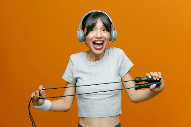 Sporty beautiful woman in sportswear with headphones on her head holding jumping rope doing exercises happy and positive standing over orange background