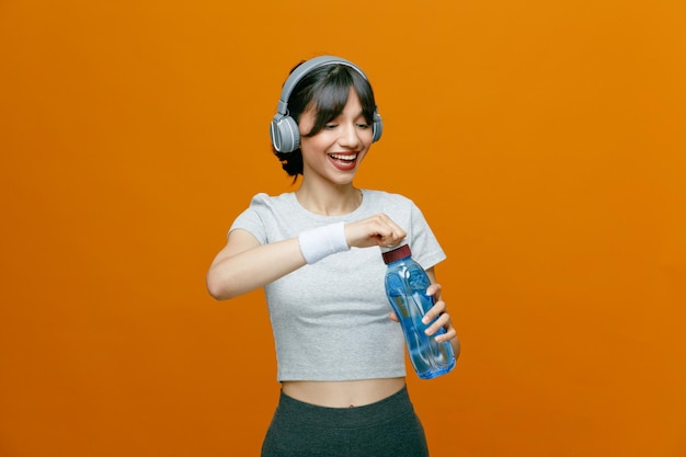 Sporty beautiful woman in sportswear with headphones holding bottle of water going to drinking smiling confident standing over orange background