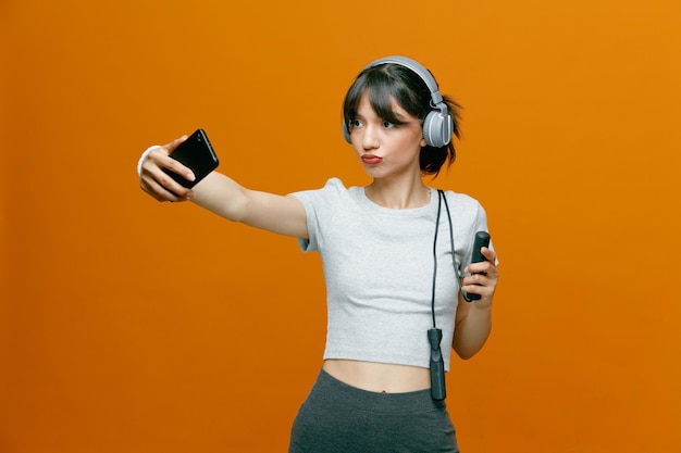 Sporty beautiful woman in sportswear with headphones using smartphone doing selfie with serious confident expression standing over orange background