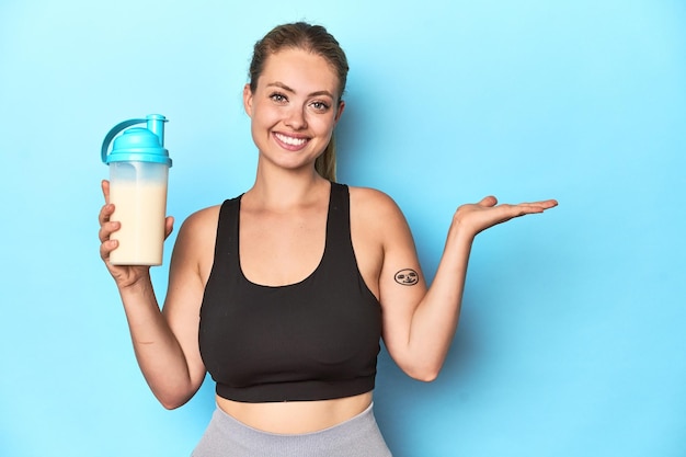 Sporty blonde with a protein shake showing a copy space on a palm and holding another hand on waist