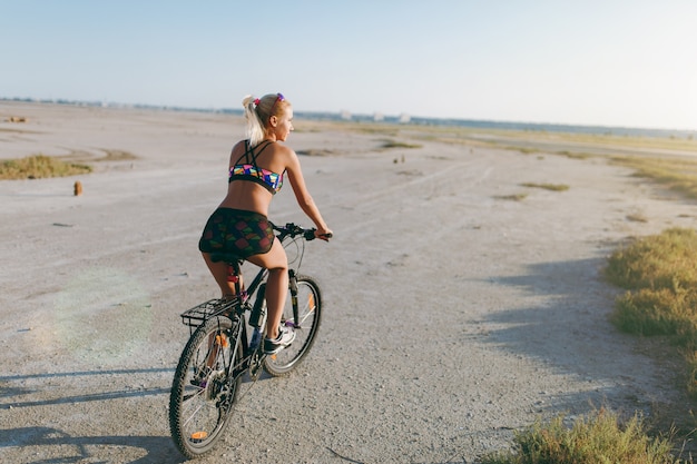 The sporty blonde woman in a colorful suit rides a bike in a desert area on a sunny summer day. Fitness concept. Back view