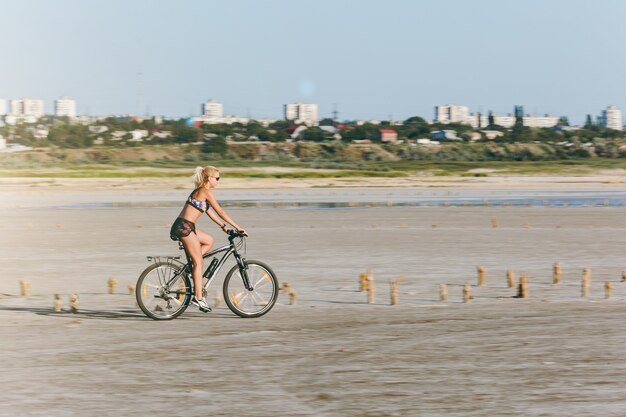 The sporty blonde woman in a colorful suit rides a bike in a desert area on a sunny summer day. Fitness concept.