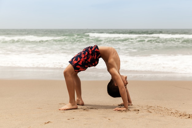 Ragazzo sportivo che fa posa sulla spiaggia, pratica della ruota di yoga di mattina all'aperto