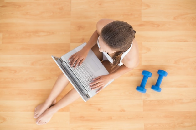 Sporty brunette using a laptop and sitting next to dumbbells