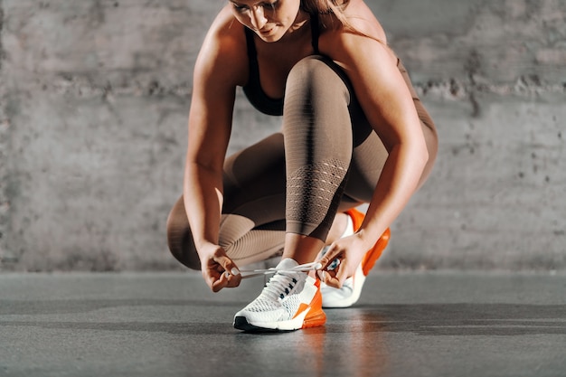 Photo sporty brunette with ponytail and in sportswear kneeling and tying shoelace in front of gray wall.