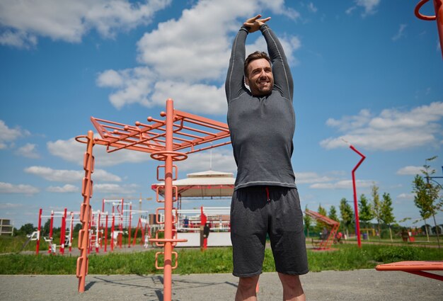 Спортивный мужчина кавказской Европы, тренирующийся на открытом воздухе. Счастливый спортсмен, наслаждаясь фитнес-тренировкой на открытом воздухе, поднимая руки вверх и выполняя разгибание и растяжку.