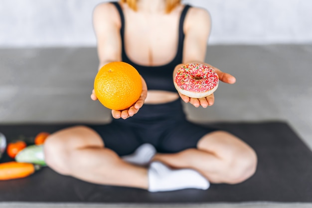 Sporty female holding donut and orange in hands. Healthy and unhealthy food concept.