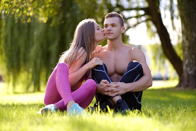 Sporty female kissing shirtless, muscular male sitting on a grass in a park.