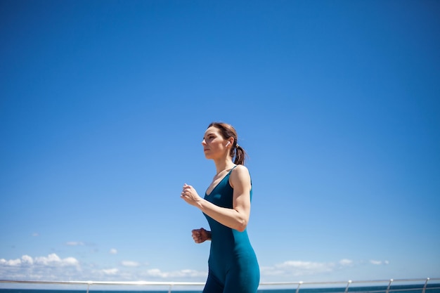 Sporty fit woman in sports clothes jogging on the embankment on a bright sunny day with blue sky and clouds