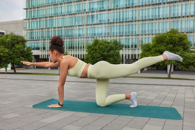 Sporty fit woman streches legs and arms does pilates exercises on fitness mat exercises every day dressed in sportswear poses against urban setting. Athletic female model involved in gymnastics