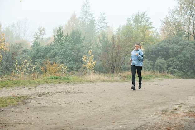 Ragazza sportiva in felpa con cappuccio blu, leggins neri e scarpe da ginnastica che fanno jogging lungo il sentiero nel bosco tra alberi verdi e gialli in una fresca mattinata nebbiosa in autunno