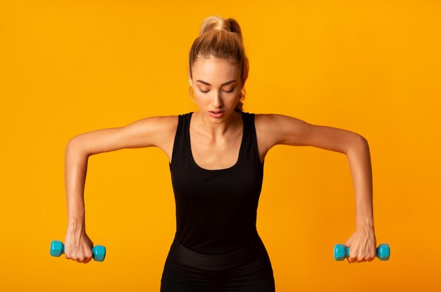 Sporty girl doing workout with dumbbells standing over yellow background