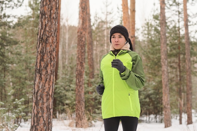 屋外でトレーニングしながら冬の森林公園で一人で走っている緑のジャケットのスポーティな女の子