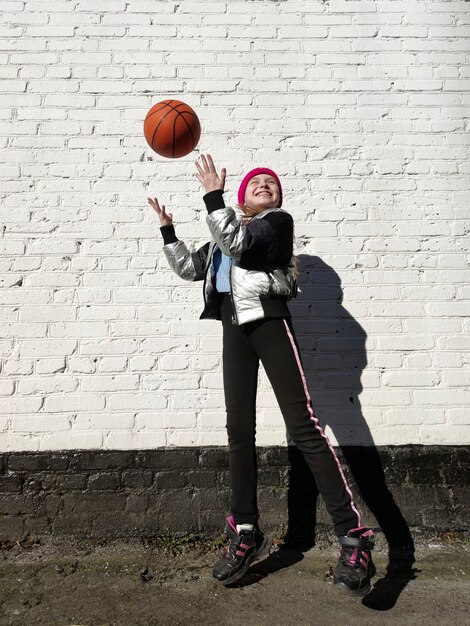 A sporty girl plays with a basketball by a white brick wall.