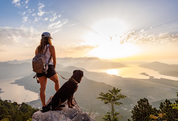 バックパックを背負ったスポーティーな女の子がロットワイラー犬と一緒に山の端に立っている