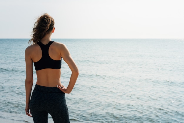 Sporty girl with curly hair in a sports bra standing on the beach and looking at the sea in the morning