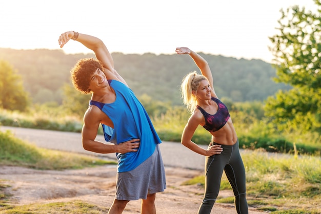 Sporty gorgeous Caucasian couple in sportswear warming up before running. Sunny summer day in nature. Three quarter length.