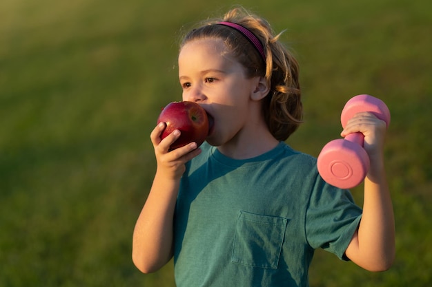 Sporty kid with apple and dumbbell outdoor in summer park Cute child boy pumping up arm muscles with dumbbell Fitness kids with dumbbells