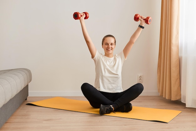 Sporty lifestyle. Young girl wearing white T-shirt and black leggins having training at home. Fit woman holding weight red dumbbells, raised arms, expressing happiness.