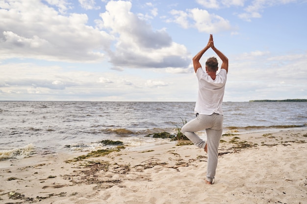 Sporty male is standing on one foot and raising hands to sky in asana on sea shore