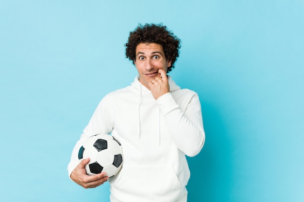 Sporty man holding a soccer ball biting fingernails, nervous and very anxious.