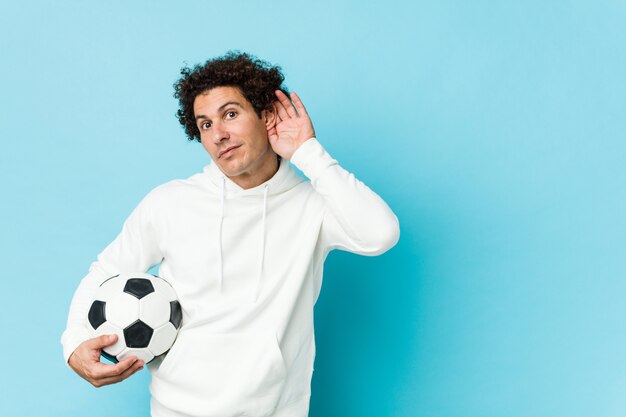 Sporty man holding a soccer ball trying to listening a gossip.