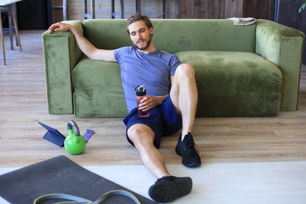 Sporty man resting, having break drinking water after doing exercise sitting on floor in living room. Fintess at home during self isolation.