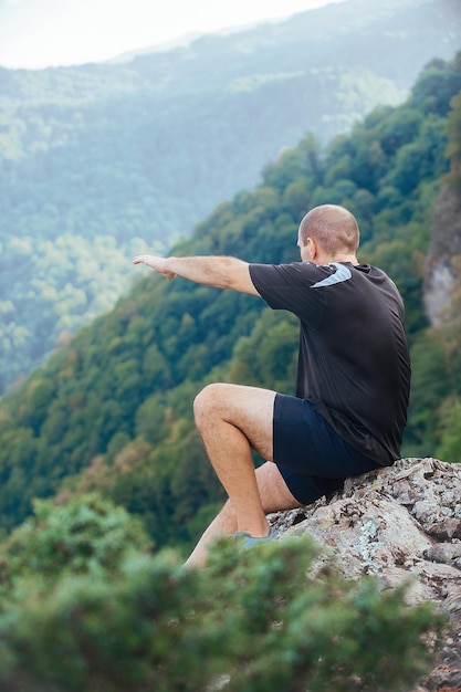 A sporty man sits on a high mountain climb during a hiking
ascent and shows his hand forward