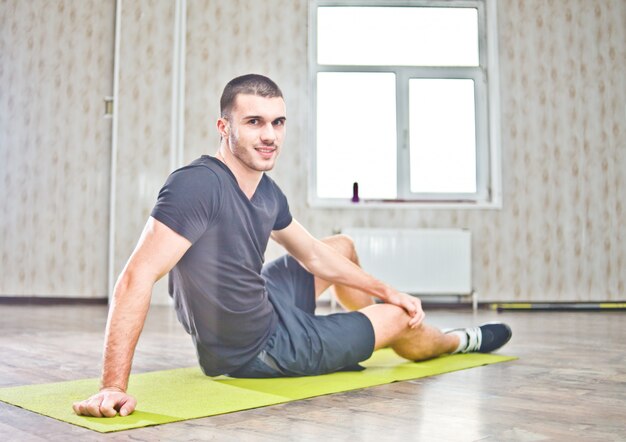 Sporty man smiling at the camera while sitting on the yoga mat and having rest after exercising.