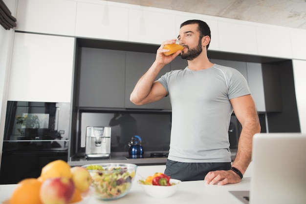 A sporty man stands in the kitchen and drinks orange juice.