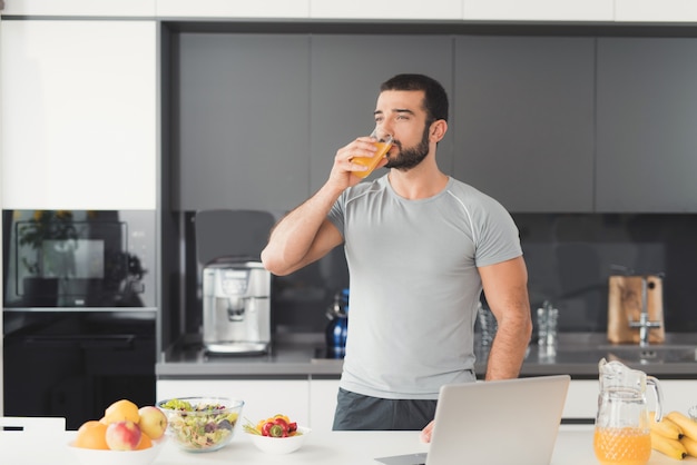 A sporty man stands in the kitchen and drinks orange juice.