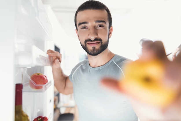 Sporty man stands in kitchen and takes fresh vegetables.